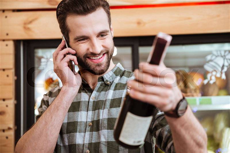 Happy young man buying wine and talking on cell phone in grocery shop, stock photo