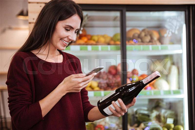 Smiling young woman choosing vine and scanning bar code on the bottle in grocery store, stock photo