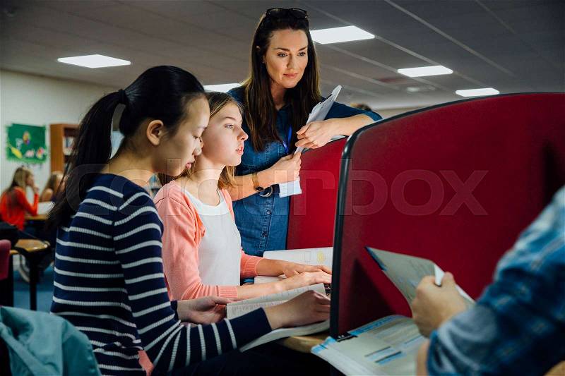 Students are working on computers with textbooks. A female teacher is standing with them, helping them with their work, stock photo
