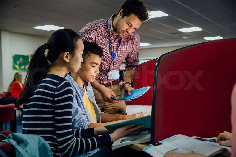 Students are working on computers with textbooks. A male teacher is standing with them, helping them with their work, stock photo