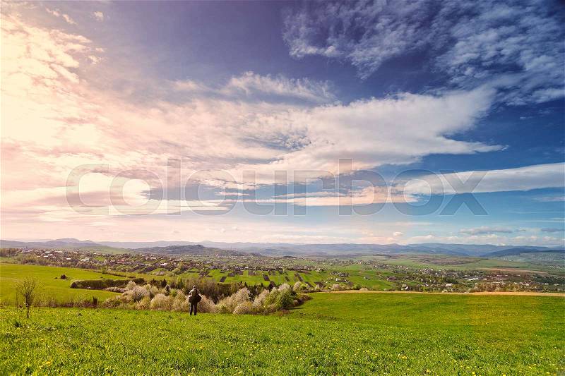 Traveler on green spring meadow. Village on sunny green spring foothills. Orchard blooming on hills. Sunny green blooming spring landscape, stock photo