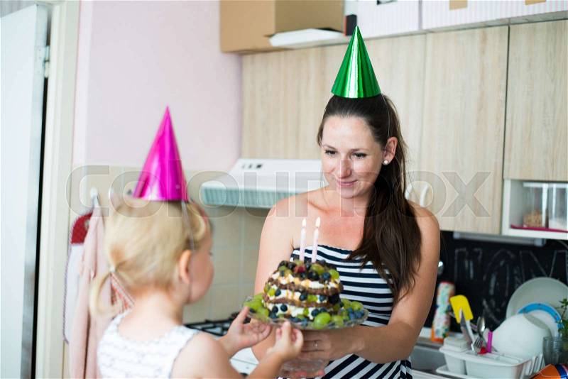 Beautiful young smiling mother giving her daughter fruit birthday cake with two candles. Colorful party hats on heads, stock photo
