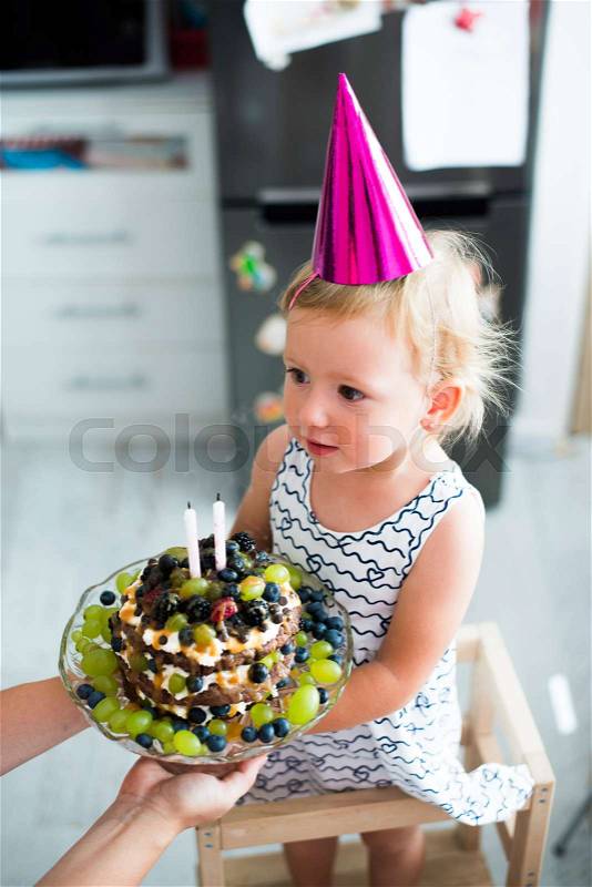 Unrecognizable young mother giving her daughter fruit birthday cake with two candles. Pink party hat on head, stock photo