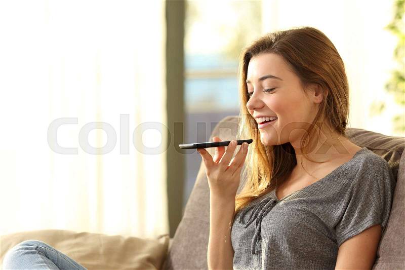Girl using a smart phone voice recognition on line sitting on a sofa in the living room at home with a warm light and a window in the background, stock photo