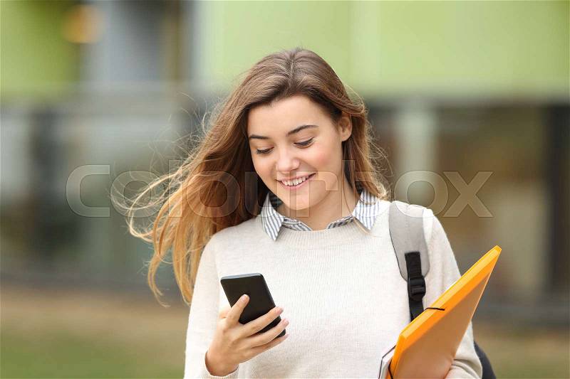 Single student walking and reading mobile phone messages with a university building in the background, stock photo