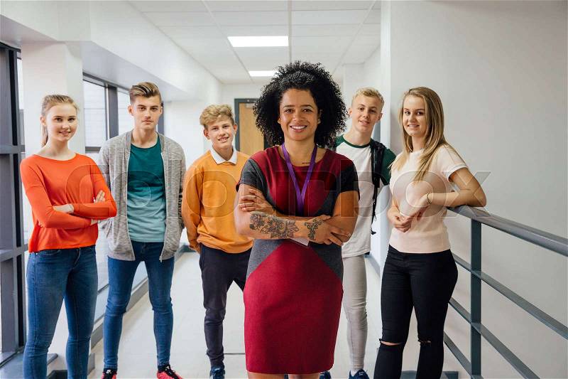 Female teacher with tattoos is posing for the camera with her arms folded. There are students in the background who are also posing for the camera, stock photo
