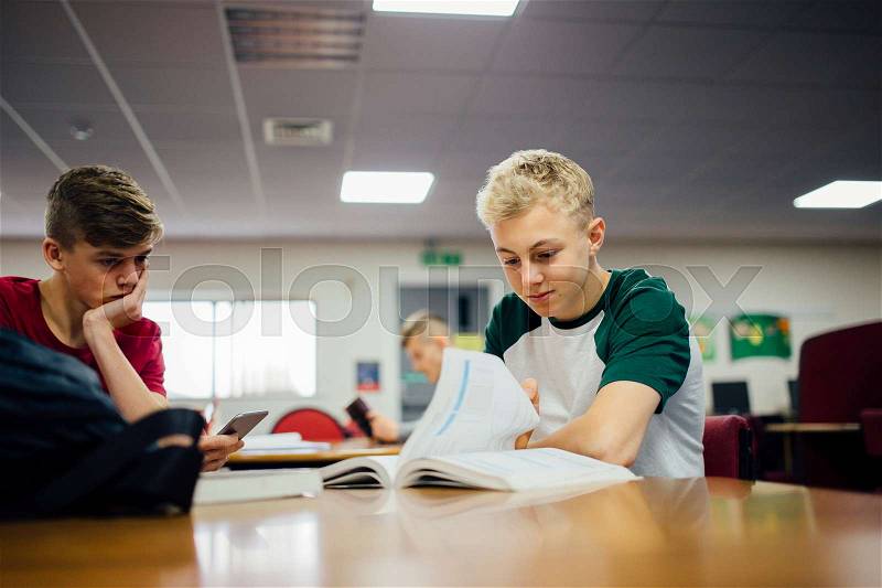 Teenage boys are at an after school club. One of the boys is catching up with work while the other is using his smart phone, looking bored of waiting for his friend. , stock photo