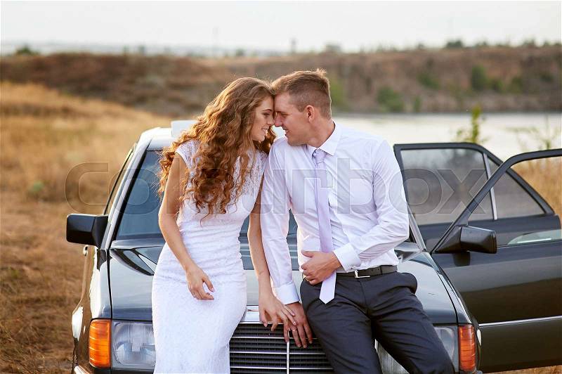 Bride and groom sit on the hood of the car and look at each other, stock photo