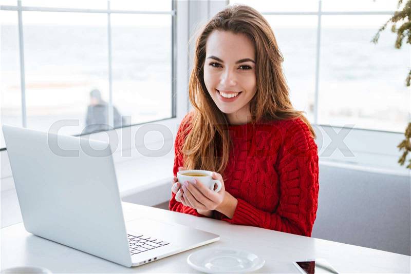 Happy cute young woman drinking coffee and working with laptop in cafe, stock photo