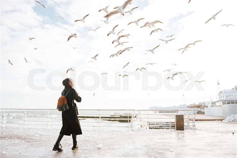 Cheerful attractive young woman walking on pier and looking at birds in winter, stock photo