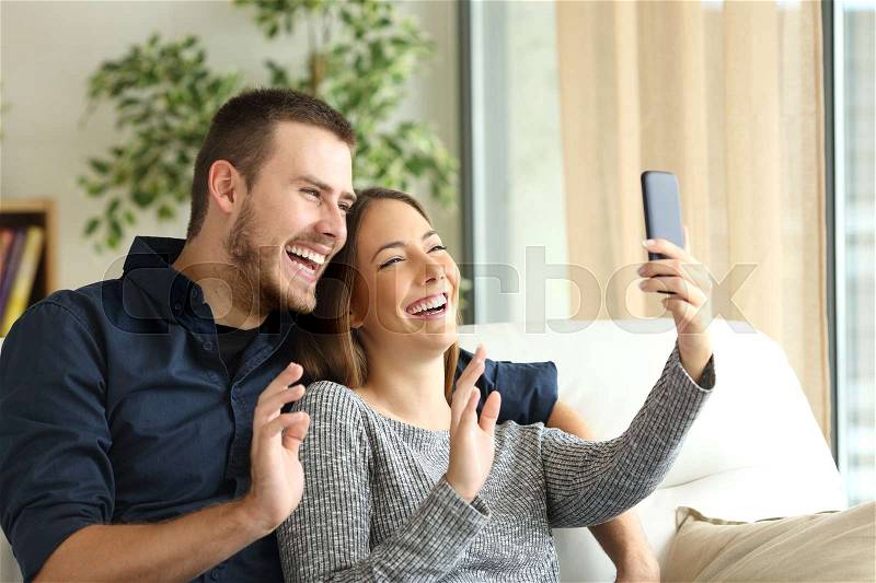 Happy casual couple greeting in a phone video call on line sitting on a sofa in the living room at home with a window in the background, stock photo
