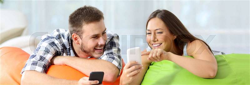 Two happy friends sharing media content on line with their smart phones lying on colorful puff in a house interior, stock photo