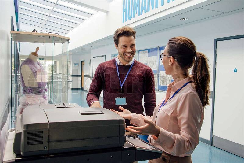 Two teachers are talking while standing at a printer in the school hall. , stock photo