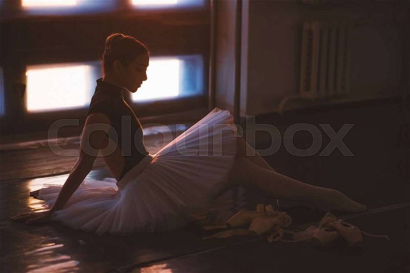 Ballerina sitting on the floor in dark ballet class against window full of sunlight, stock photo
