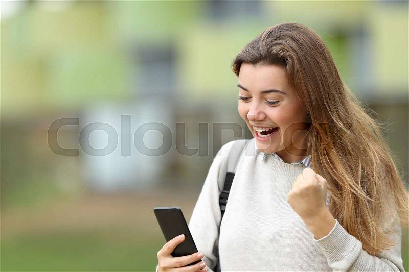 Excited student reading good news on line in a smart phone in the street with the university building in the background, stock photo