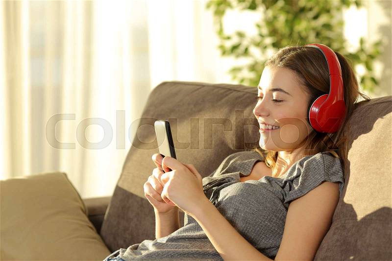 Single teen listening music on line with a smart phone and headphones sitting on a sofa n the living room at home, stock photo