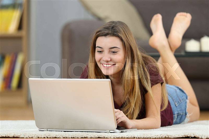 Happy teenager girl on line with a laptop lying on a carpet on the floor in the living room at home, stock photo