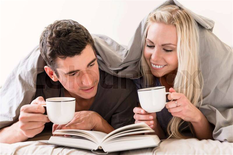 Young happy couple drinking coffee while reading a book together in bed, stock photo