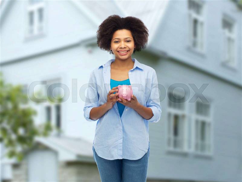 People, investment, saving and finances concept - happy afro american young woman with piggy bank over house background, stock photo