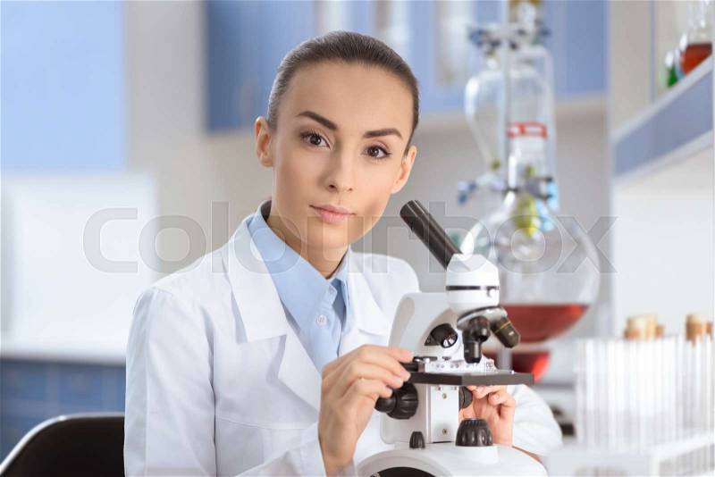 Young woman scientist in lab coat working with microscope and looking at camera, stock photo