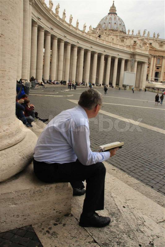 The solitary believer is sitting against a collumn and has an open bible in his hands and is looking to the people on the St. Peter's Square in the city Rome in the summer, stock photo