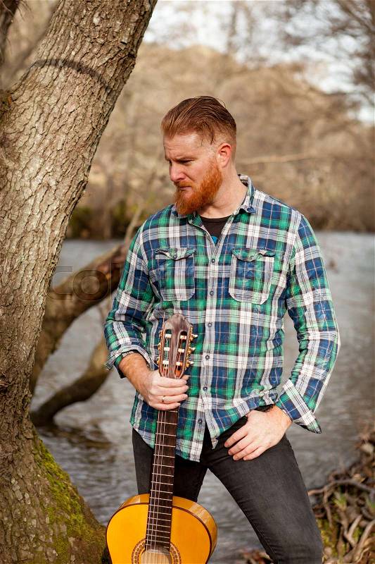 Hipster man with red beard holding a guitar in the field, stock photo