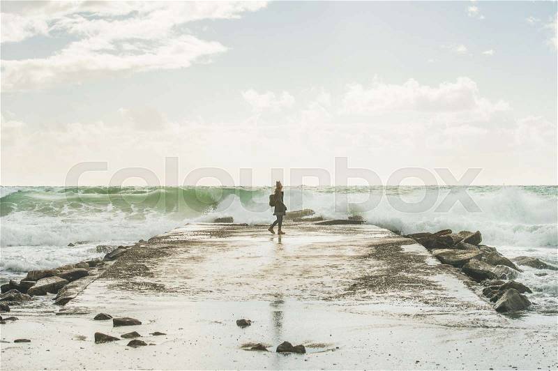 Silhouette of young woman walking on pier surrounded by stones and looking at waves of stormy Mediterranean sea in winter, Alanya, Turlkey, stock photo