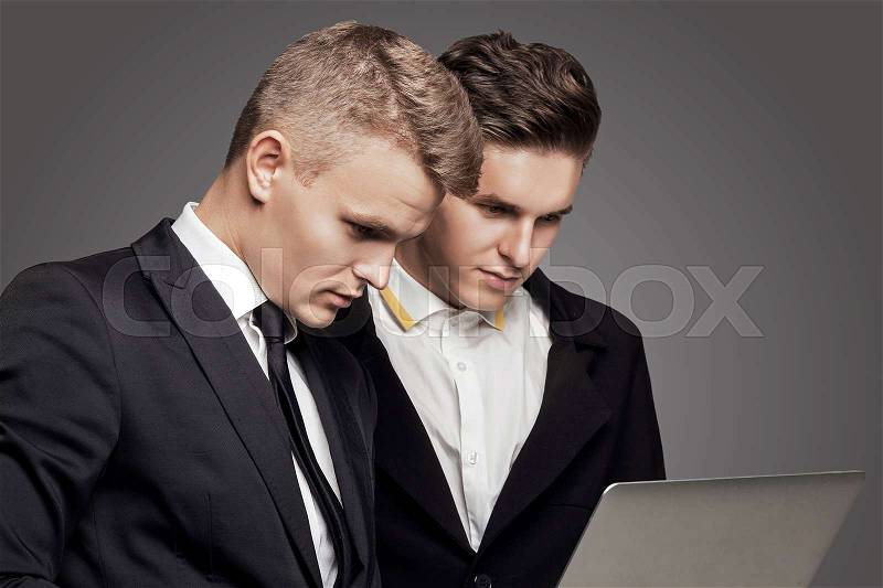 Two young elegant mans lookking on laptop. Black suite, gray background. Studio, stock photo