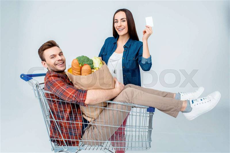 Happy man sitting in shopping cart with grocery bag and smiling woman showing blank card, stock photo