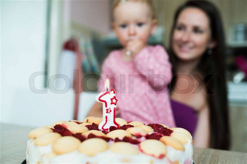 Beautiful young smiling mother with her cute daughter celebrating her first birthday. Birthday cake with candle laid on table, stock photo