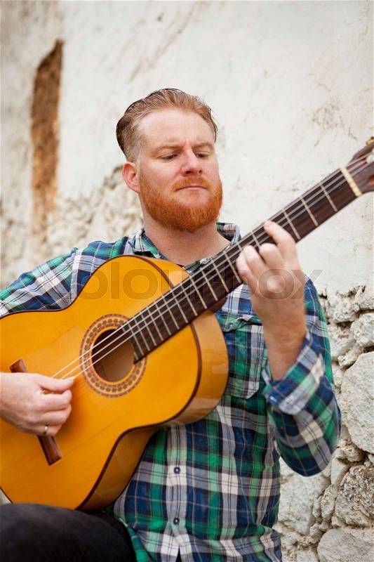 Hipster man with red beard playing a guitar with a old wall of background, stock photo