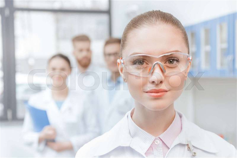 Close-up portrait of young woman scientist in lab coat and protective goggles smiling, stock photo