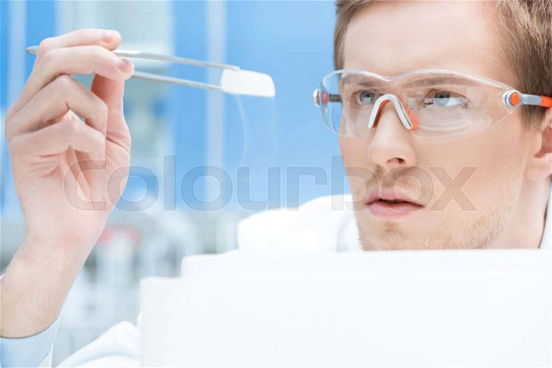 Concentrated scientist in protective glasses and gloves looking at chemical sample, stock photo