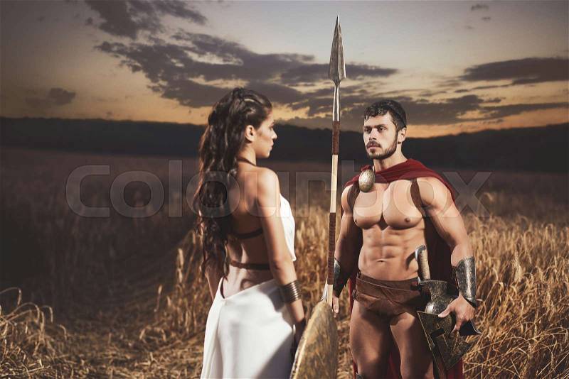 Portrait of brunette woman wearing like greece in white dress and man warrior wearing like spartan. Couple meeting after war in field at night time, stock photo
