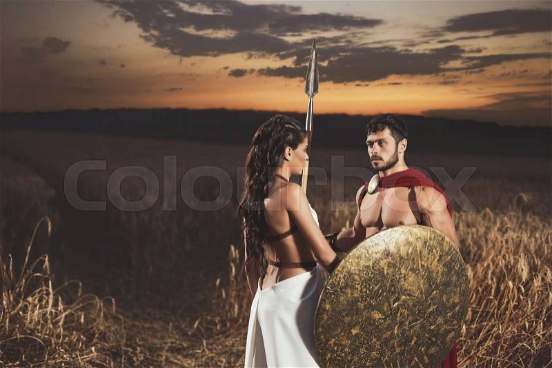 Portrait of couple meeting after war in field. Beautiful woman with long hair wearing like greece in white dress looking face to face with man warrior wearing like spartan, stock photo