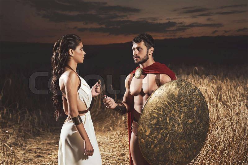 Portrait of brunette woman wearing like greece in white dress giving amulet to man with bread, warrior wearing like spartan. Couple meeting after war at night time, stock photo