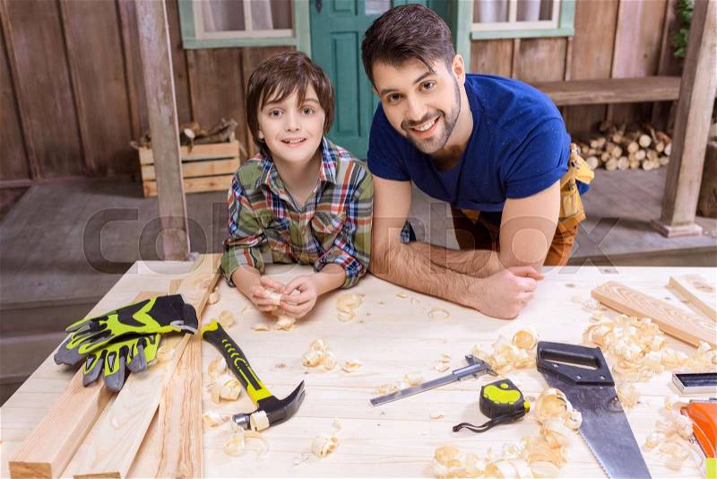 Happy father and son leaning on table with tools and smiling at camera, stock photo