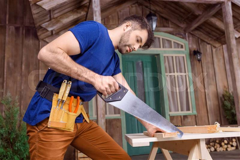 Low angle view of handsome bearded carpenter in tool belt sawing wooden plank on porch , stock photo
