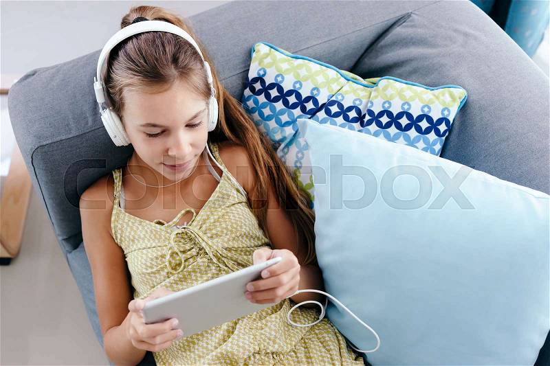 10 years old tween girl relaxing on a couch, listening to music in headphones and playing with tablet pc. Child chilling on the sofa in living room, stock photo