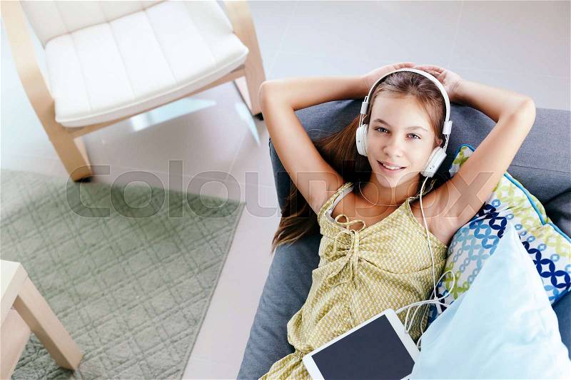 10 years old tween girl relaxing on a couch, listening to music in headphones and playing with tablet pc. Child chilling on the sofa in living room. View from above, stock photo