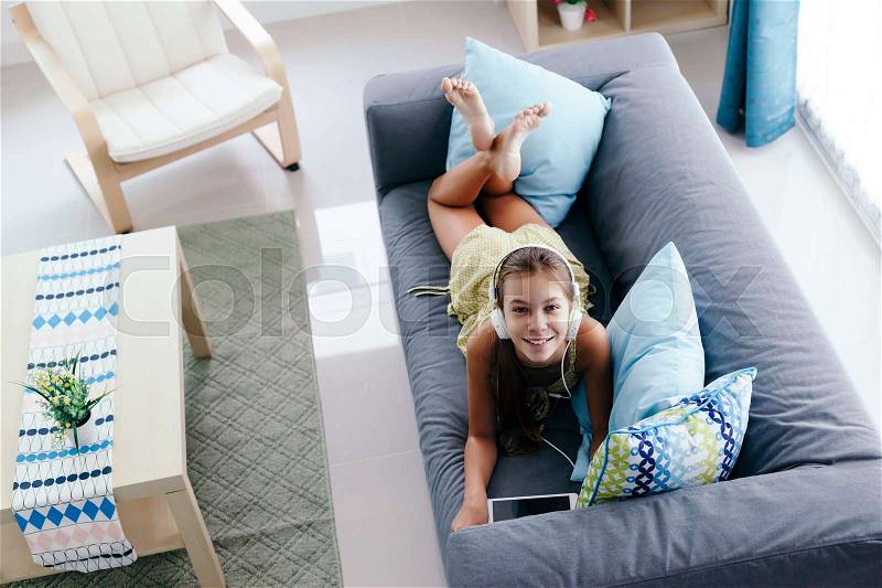 10 years old tween girl relaxing on a couch, listening to music in headphones and playing with tablet pc. Child chilling on the sofa in living room, stock photo