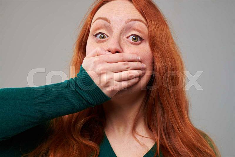 Close-up hot of scared young red hair woman covering her mouth with hand. Female with freckles isolated over gray background in the studio , stock photo