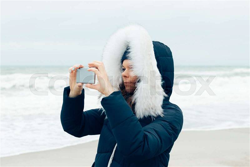 Young woman in a winter jacket with a hood using a mobile phone on the beach, close-up, stock photo