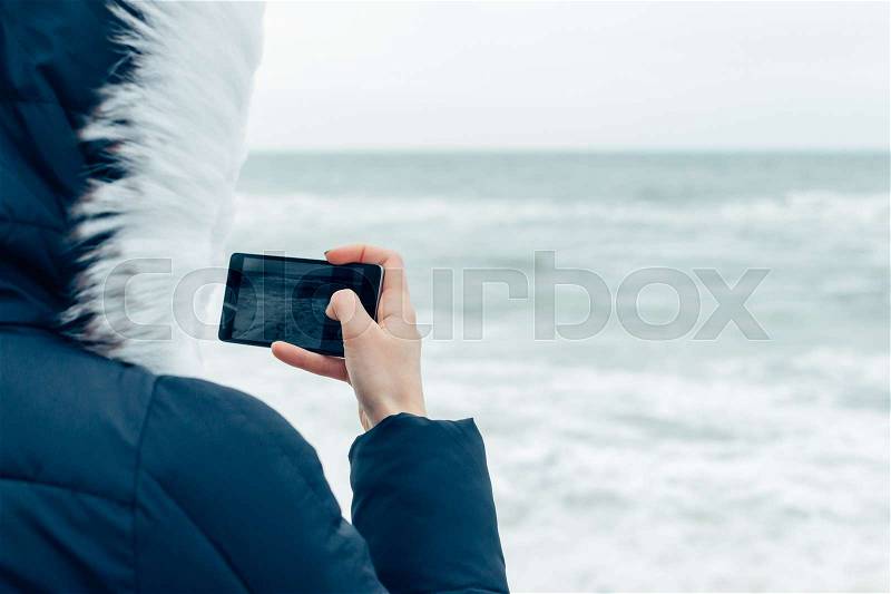 Close-up of a woman in a winter jacket with a hood using a mobile phone on the beach, stock photo