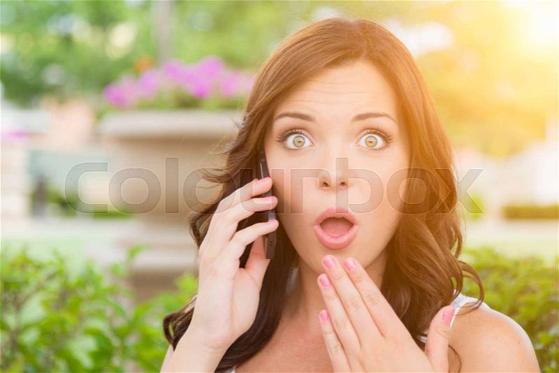 Shocked Young Adult Female Talking on Cell Phone Outdoors on Bench, stock photo