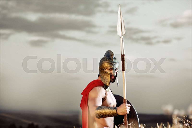 Empire warrior. Ancient shirtless warrior with perfect strong muscular body wearing a helmet and a cape standing in the field with a shield and a spear under grey sky copyspace confidence concept, stock photo