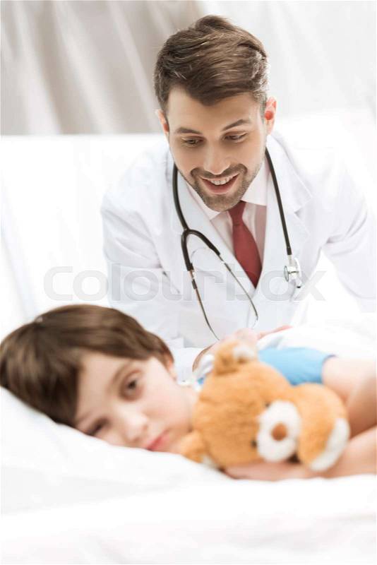 Upset child patient lying in bed with smiling doctor behind, stock photo