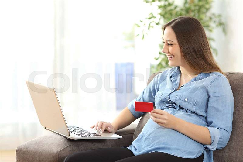 Pregnant woman buying on line with credit card and a laptop sitting on a sofa in the living room at home, stock photo