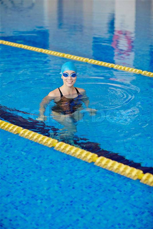 Fit young woman wearing cap and goggles standing in clear blue water of swimming pool between yellow lines, looking at camera and smiling, stock photo