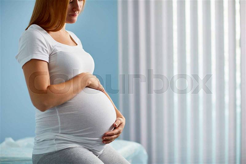 Side view image of woman with big belly in her last months of pregnancy sitting on bed in doctors office against window waiting for her examination, stock photo